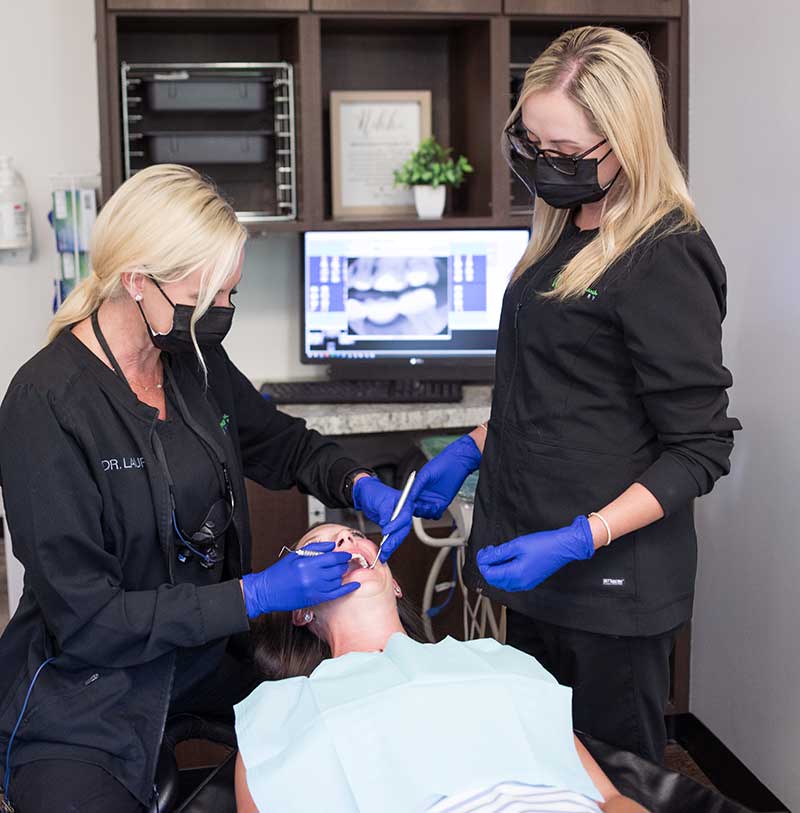 dentist and her assistant looking in a patients mouth in an office with the image of a tooth X-ray on the computer